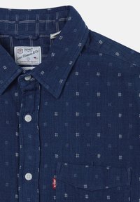 Camicia in jeans blu navy con motivi geometrici bianchi, chiusura con bottoni, colletto, taschino singolo sul petto e etichetta rossa vicino alla cucitura della tasca.
