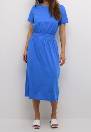 Nainen, jolla on päällään keskipitkä, lyhythihainen kuninkaansininen mekko, jossa on joustava vyötärö, ja valkoiset avonaiset liukuvasteet.