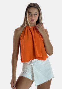 Top a halter arancione dal design fluido, abbinato a pantaloncini di jeans bianchi con taglio asimmetrico e orli sfrangiati.