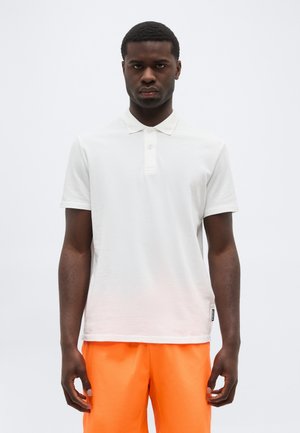 Egy férfi, aki egyszerű fehér rövid ujjú galléros pólót és élénk narancssárga rövidnadrágot visel, semleges világos háttér előtt áll.