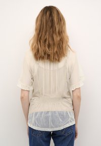 Gennemsigtig cremet blouse med smocked-detaljer bagpå, korte flæseærmer og en prikket tekstur. Bæres med blå jeans.