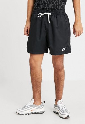 Mand iført sorte Nike-shorts med hvid snøre og hvide Nike-sneakers mod en ensfarvet baggrund.