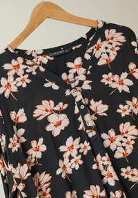 Blusa nera con un motivo floreale in crema e arancione, scollatura a V, tre bottoni e vestibilità rilassata.