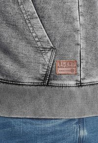 Grijs sweatshirt van gestructureerde stof met geribbelde manchetten en zoom. Heeft een bruine leren patch met het opschrift "BLEND" en de tekst "GLOBAL JEANMAKER."