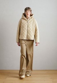 Ελαφριά μπεζ μπουφάν με κουκούλα, με φερμουάρ μπροστά και διαγώνιες ραφές· συνδυασμένο με καφέ παντελόνι φαρδύ και ανοιχτόχρωμα παπούτσια.