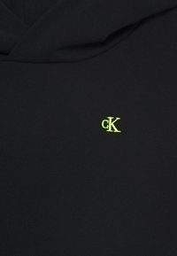 Schwarzer Hoodie-Stoff mit kleinem, neon-grünem "cK"-Logo, das auf der Brust in der Nähe des Ausschnitts gestickt ist.