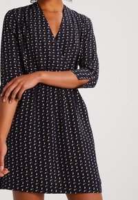 Robe marine avec un décolleté en V, des manches trois-quarts, et un motif présentant de petites formes jaunes et blanches partout. Tissu doux.