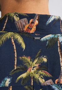 Camicia a maniche corte con sfondo blu navy e motivo di palme, chitarra acustica e disegno di batteria. Il tessuto appare morbido e leggero.