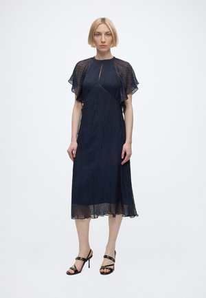 Frau mit kurzen blonden Haaren, die ein marineblaues Midikleid mit transparenten, flatternden Ärmeln und schwarzen, dünnen High Heels trägt, steht vor einem einfarbigen Hintergrund.