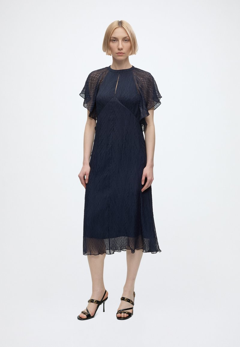 Femme aux cheveux blonds courts portant une robe midi bleu marine avec des manches flottantes transparentes et des escarpins noirs à fines lanières, debout devant un fond uni.