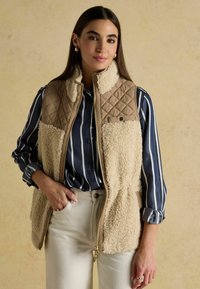 Beež ja kreemjas fliisvest, millel on tikkimine pruunis ülaosas, ees lukuga ja väike roheline aktsent. Kombineeritud tumesinise triibulise särgiga.