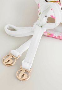 Bretele din material textil alb legate în nod, cu capete metalice circulare aurii embossate cu o literă „b” stilizată, pe o suprafață deschisă la culoare, lângă material floral.