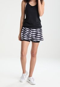 Svart linne ihop med svart och vitt mönstrade shorts med vinklade mönster. Modell som bär vita träningsskor.