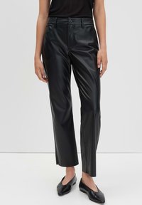 Vrouw in zwarte faux leren rechte broek met knoop en rits, gecombineerd met zwarte puntige ballerina's en een zwart topje.