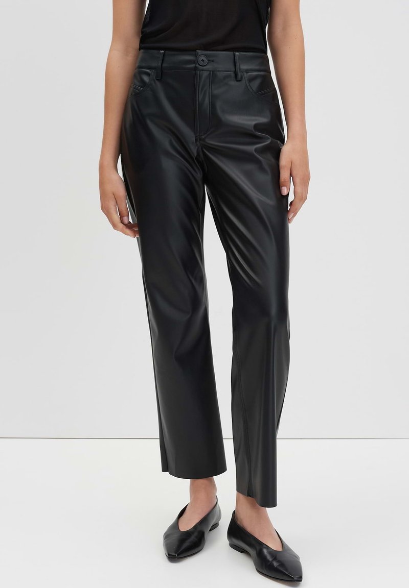 Vrouw in zwarte faux leren rechte broek met knoop en rits, gecombineerd met zwarte puntige ballerina's en een zwart topje.