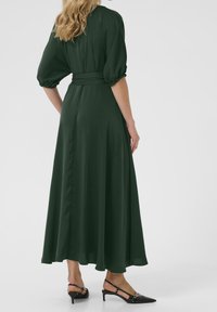 Groene, vloeiende maxi-jurk met korte pofmouwen en een gebonden taille. Gemaakt van een gladde stof met een volle rok; gecombineerd met zwarte puntige hakken.