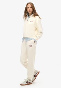 Crème fleece pullover met een kwart rits en logo, gecombineerd met crème joggers met logo patch. Sneakers met mintgroene en bordeauxrode accenten.