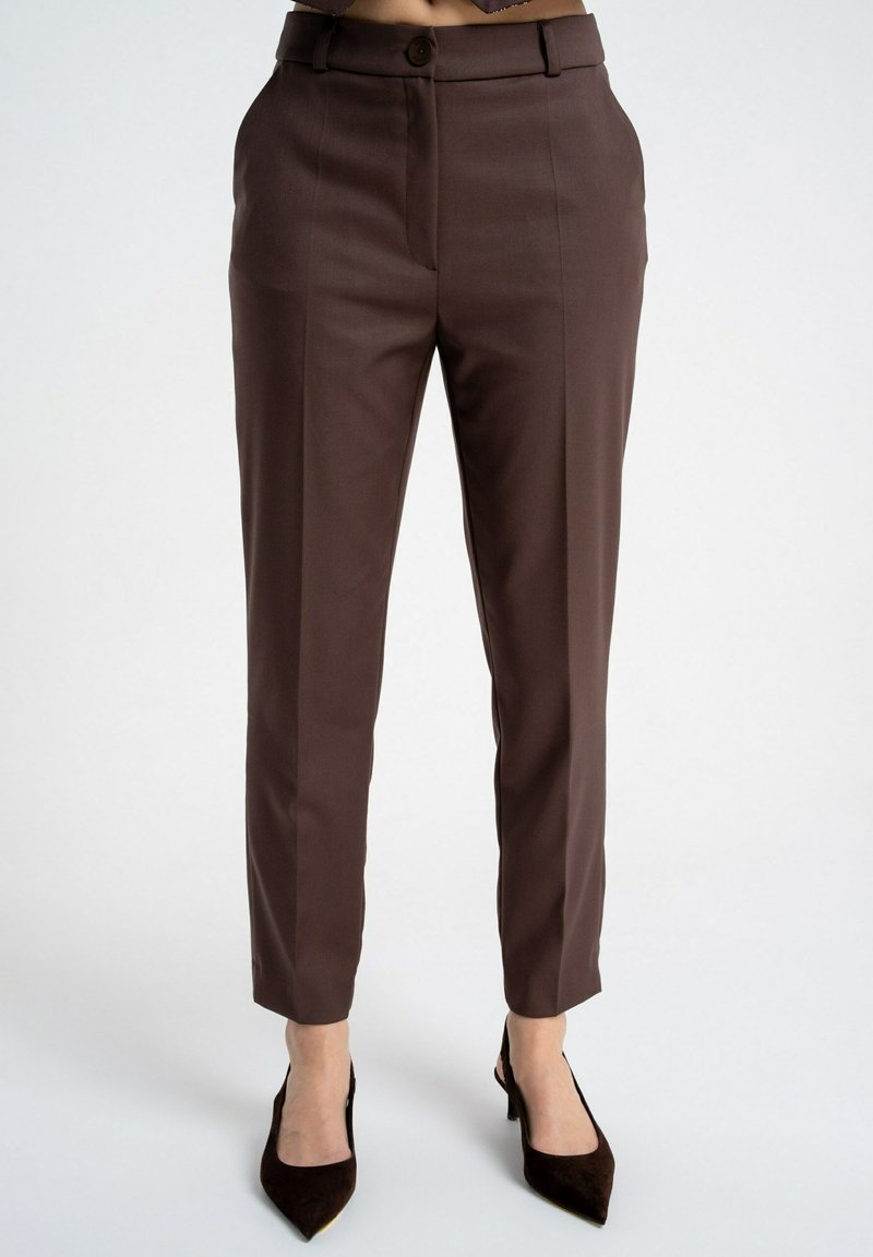 Brązowe, dopasowane spodnie z wysokim stanem, z przednią plisą, szlufkami na pasek i zapinane na guzik, noszone z czarnymi szpilkami z ostrym noskiem na białym tle.