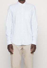 Jasnoniebieska koszula zapinana na guziki w pionowe białe paski, z kieszonką po lewej stronie klatki piersiowej i długimi rękawami. Materiał wydaje się gładki i lekki.