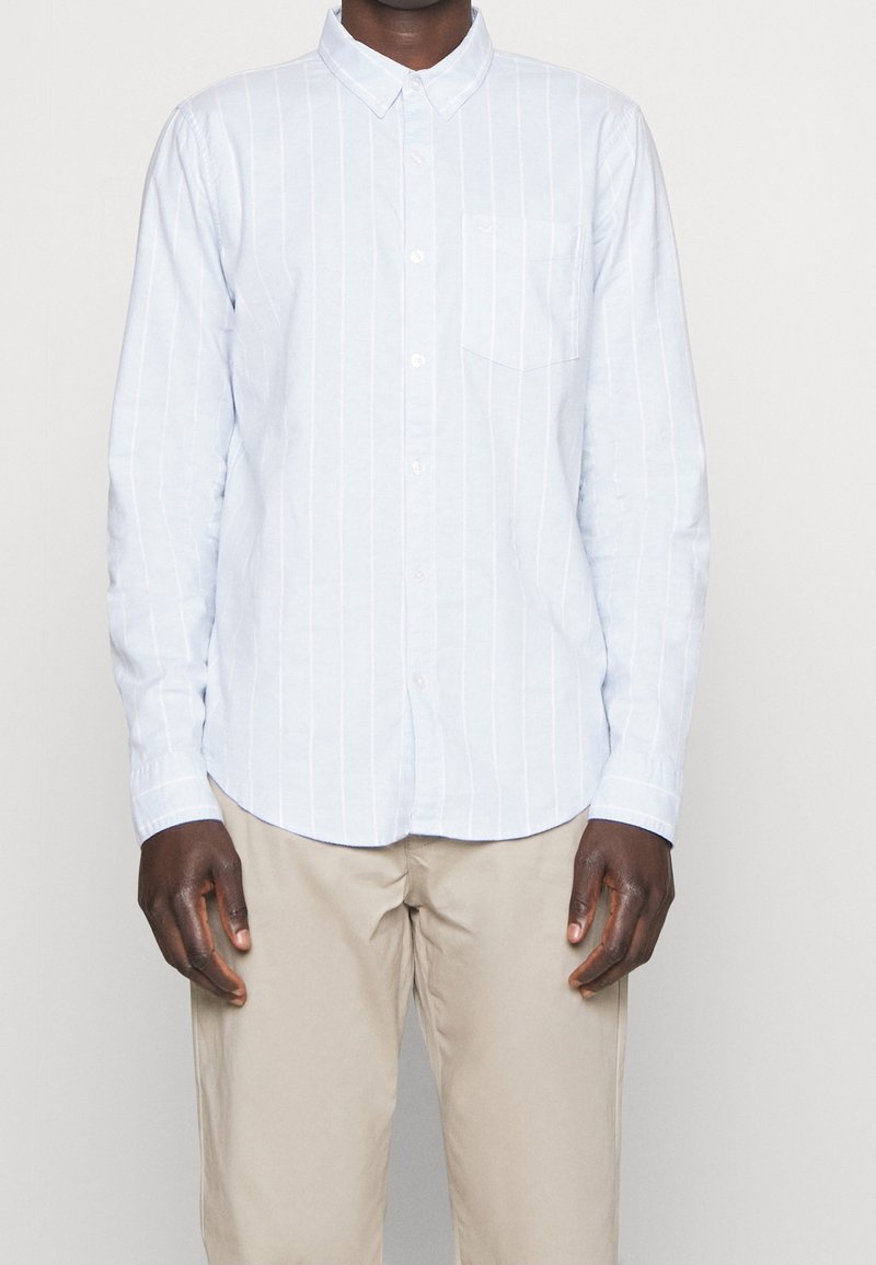 Jasnoniebieska koszula zapinana na guziki w pionowe białe paski, z kieszonką po lewej stronie klatki piersiowej i długimi rękawami. Materiał wydaje się gładki i lekki.