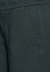 Zbliżenie na ciemnozielone spodnie z materiału, pokazujące boczną kieszeń oraz szczegółowe przeszycia na pasie i szwach.