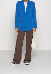 Blue tailored blazer with notched lapels, paired with brown wide-leg trousers. White t-shirt underneath and a textured white bag. Black sneakers.