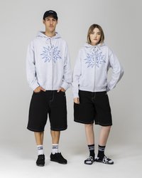 Graue Kapuzenpullis mit blauen Sonnenmotiven, kombiniert mit schwarzen Shorts. Das männliche Model trägt schwarze Schuhe, während das weibliche Model Sandalen und gemusterte Socken trägt.
