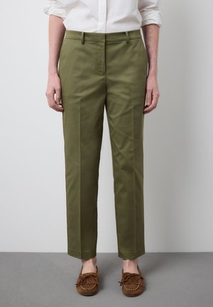 Persona che indossa pantaloni su misura verde oliva e mocassini marroni, in piedi contro una parete e un pavimento grigio chiaro.