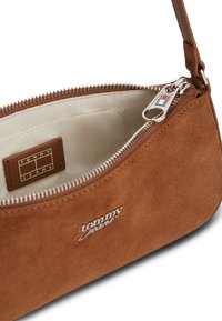 Μικρή τσάντα χειρός από καφέ σουέντ με ασημί φερμουάρ, μπεζ εσωτερικό και λογότυπο Tommy Jeans μπροστά και στην ετικέτα στο εσωτερικό.