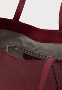 Hlboká červená textúrovaná kožená taška s béžovým látkovým interiérom, viditeľná zipsová vrecko a dva ramenné popruhy.