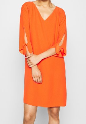 Femme portant une robe orange vif jusqu'aux genoux avec un col en V et des manches mi-longues avec des fentes nouées.