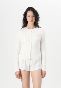 Hvit kabelstrikket cardigan med rund hals og knappelukking. Har ribbestrikket mansjetter og kant. Diskret teksturert mønster.