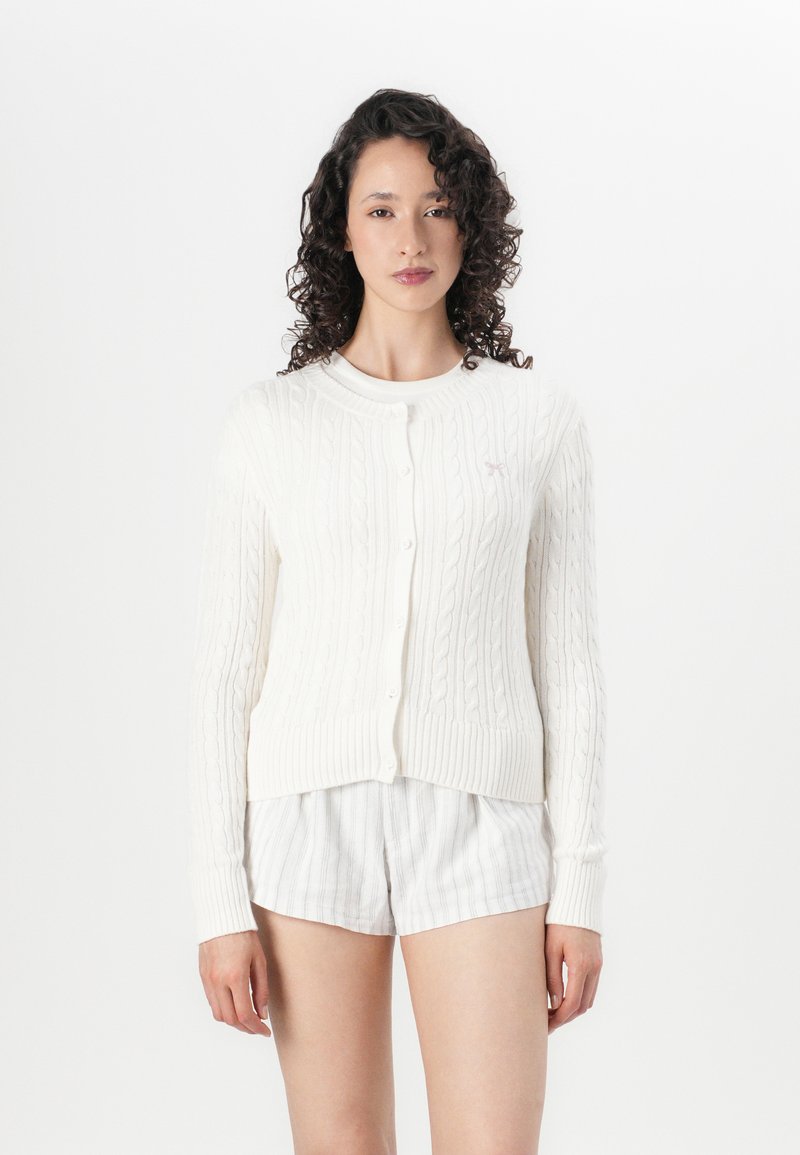 Hvit kabelstrikket cardigan med rund hals og knappelukking. Har ribbestrikket mansjetter og kant. Diskret teksturert mønster.
