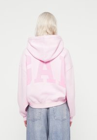 Sudadera rosa con un gran logotipo "GA" en un tono rosa claro en la parte de atrás. El diseño presenta un corte holgado y está hecha de una tela suave y gruesa.