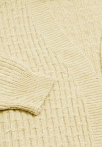Maglione lavorato a maglia color crema con motivo testurizzato e polsini e colletto a coste, piegato e appoggiato piatto.