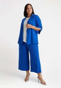 Niebieski garnitur w kolorze kobaltowym, składający się z dopasowanej marynarki z podwiniętymi rękawami oraz szerokich spodni, zestawiony z białą bluzką i srebrnymi sandałami na obcasie.