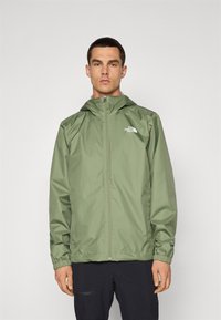 Veste à capuche verte en matière légère et résistante à l'eau, avec des poignets élastiques et une fermeture éclair à l'avant, arborant un logo sur la poitrine.