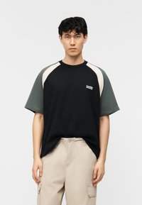 Ung mann med kort svart hår iført en t-skjorte i svart og mørkegrønn fargeblokking og beige cargobukser mot en ensfarget hvit bakgrunn.