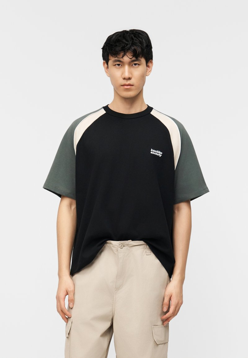 Ung mann med kort svart hår iført en t-skjorte i svart og mørkegrønn fargeblokking og beige cargobukser mot en ensfarget hvit bakgrunn.