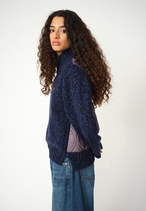 Gebreide trui in marineblauw met een textuurpatroon, hoge kraag en gewatteerde schouderaccenten. Gekocht met lichtblauwe rechte jeans.
