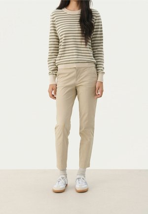 Personne portant un haut à manches longues beige rayé, un pantalon beige, des chaussettes blanches et des baskets blanches, debout devant un fond clair uniforme.