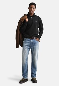 Sweatshirt preta com fecho de correr, jeans azul claro, casaco castanho pendurado no braço, sapatos escuros com detalhes dourados. Textura lisa, ajuste casual.