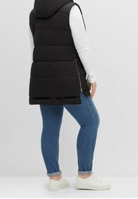 Schwarze gepolsterte Weste mit Kapuze, seitlichem Reißverschlussdetail, über einem weißen Langarmoberteil getragen, kombiniert mit blauen Jeans und weißen Turnschuhen.