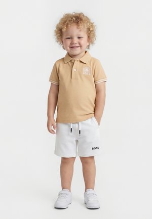 Niño pequeño con cabello rizado y rubio, vistiendo un polo beige y pantalones cortos blancos, de pie con una mano en el bolsillo, sonriendo.