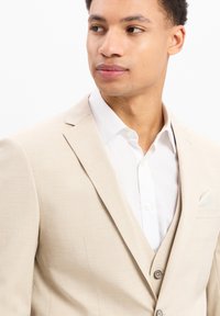 Beige colbert met een gladde textuur, voorzien van doorlopende revers, twee knopen en een kleine pochet. Witte overhemd eronder.