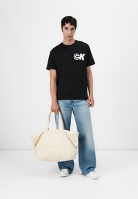 Camiseta preta com logotipo branco, calças de ganga largas em azul claro, segurando uma grande tote bag bege com logotipo em relevo e alças duplas.