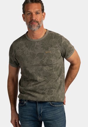 Middelbare man met grijze baard, die een olijfgroen T-shirt met tropische print en een blauwe spijkerbroek draagt, staand tegen een effen lichte achtergrond.