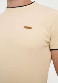 Chemise beige texturée à manches courtes avec bordure noire au col et aux manches, avec un petit patch marron portant l'inscription "PIER ONE" sur la poitrine.