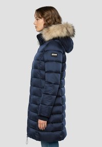 Manteau matelassé bleu marine avec capuche bordée de fourrure, présentant un design matelassé, des poignets côtelés et un logo de marque sur la poitrine. Vue de côté.