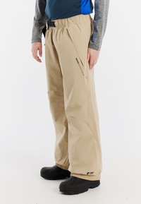 Beige waterdichte broek met een elastische tailleband en een verstelbare zwarte riem. Heeft een losse pasvorm en zwarte accenten bij de zoom en het logo.